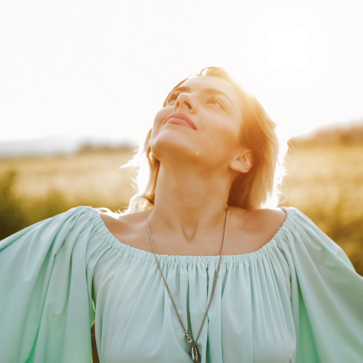 Woman outdoors with eyes closed, tilting her head back toward the sun, conveying calm, relaxation, and nervous system balance in a natural setting.