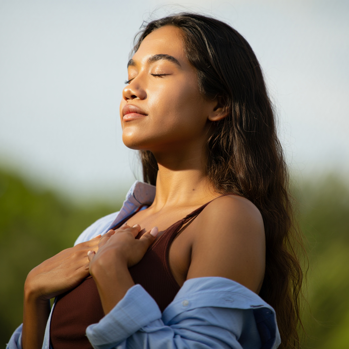 Woman standing outdoors with her eyes closed and hands on her chest, practicing a calming breathing exercise for stress relief.