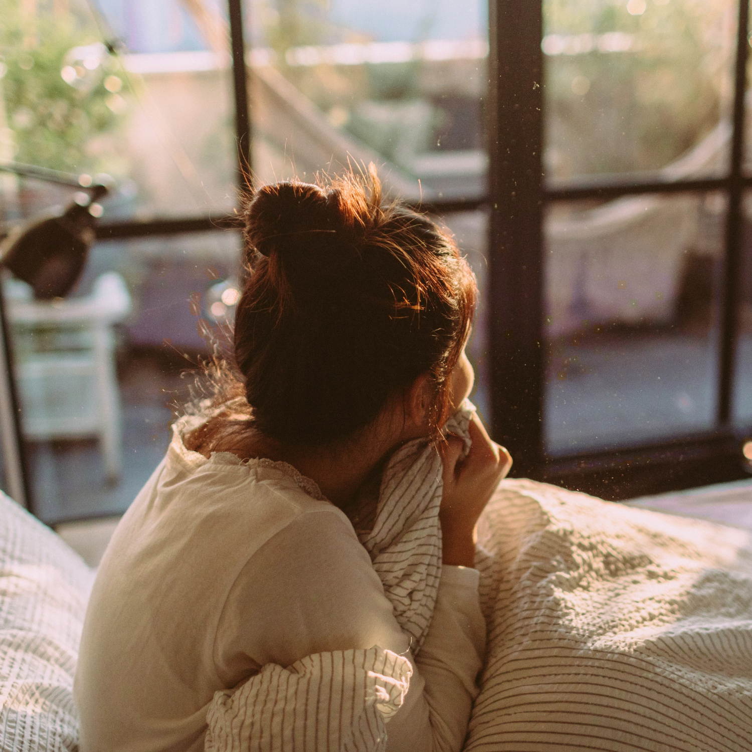Woman sitting by a sunlit window in the morning, wrapped in a cozy sweater, looking outside — evoking calm and intention at the start of the day.