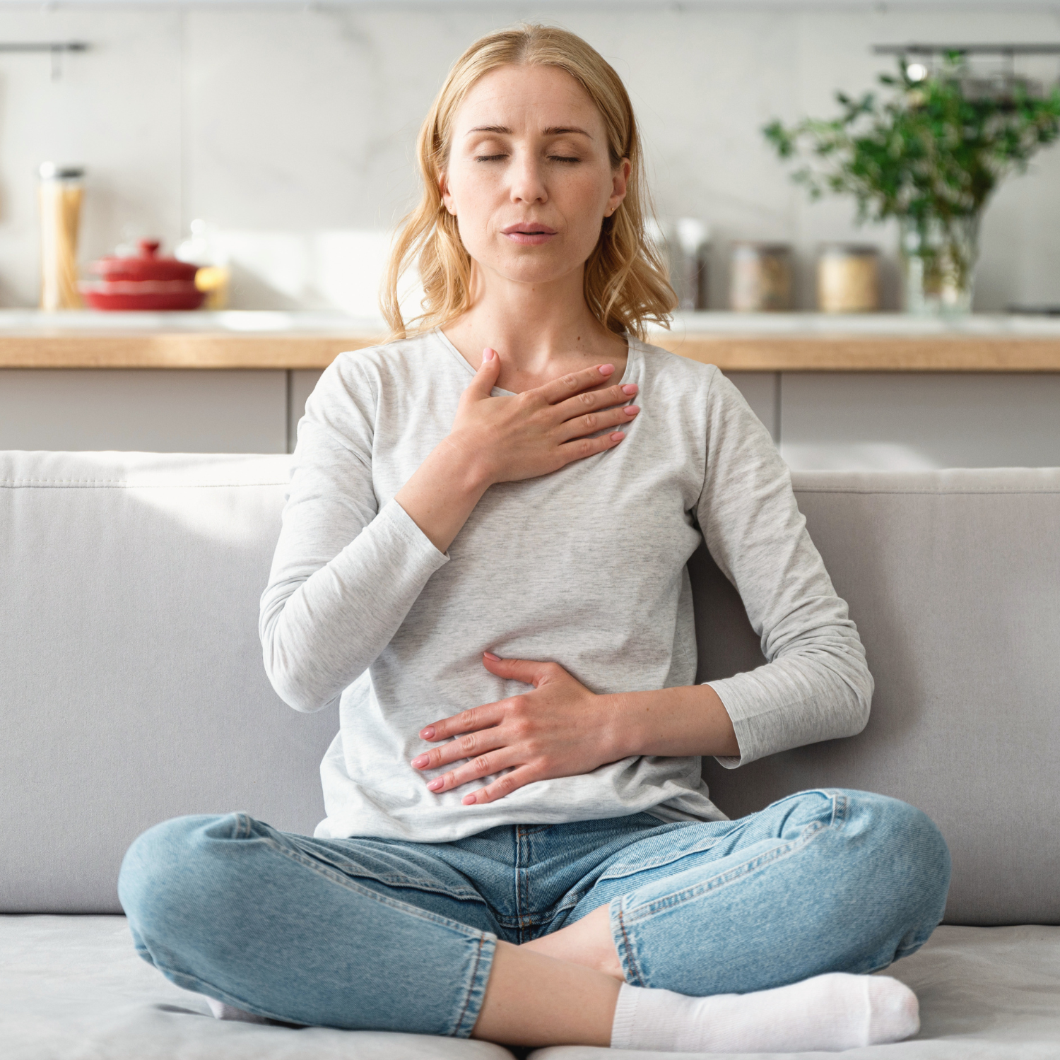 Woman sitting cross-legged on a couch with eyes closed, one hand on her chest and one on her belly, practicing diaphragmatic breathing for nervous system calm.