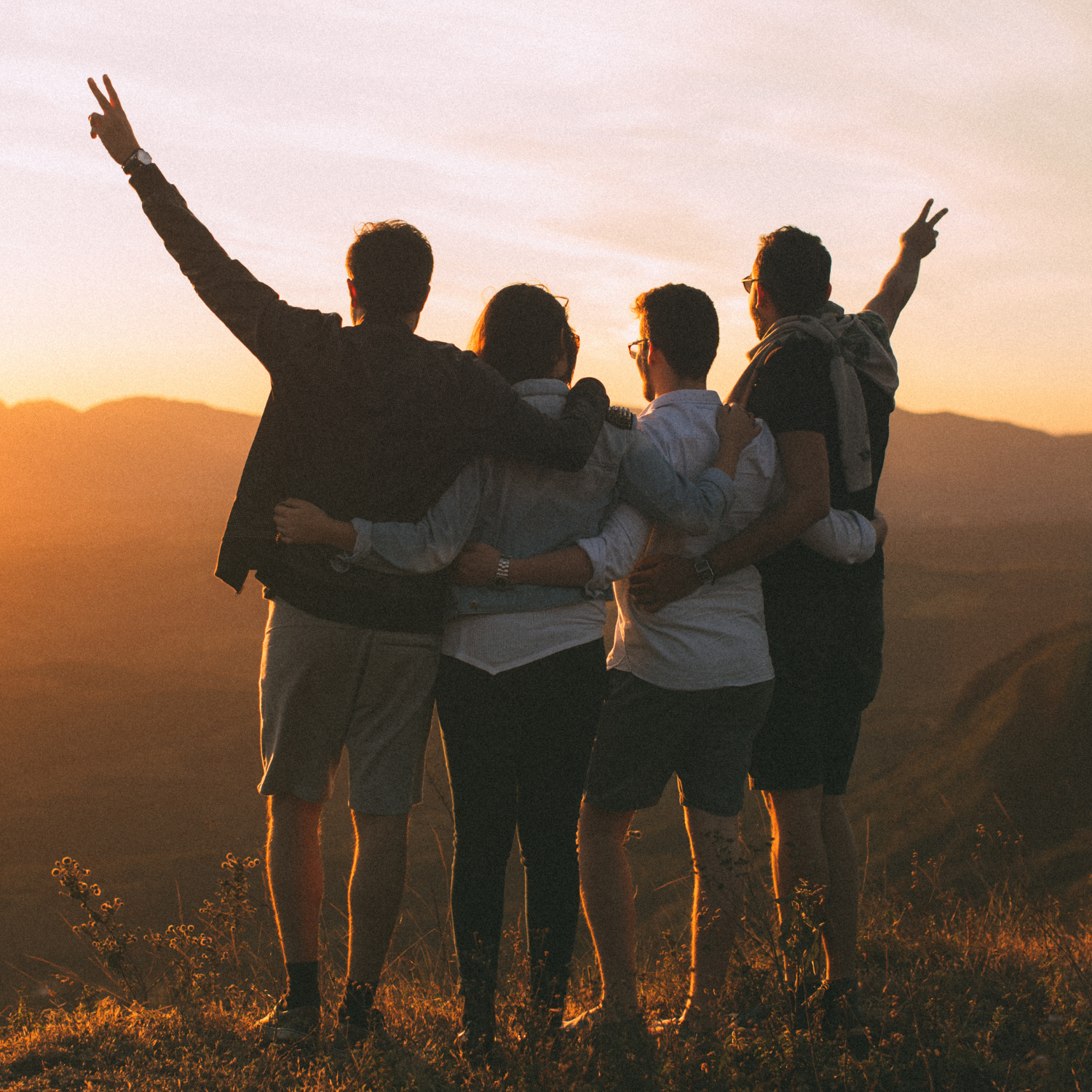 A group of four friends stands arm in arm on a hilltop at sunset, looking out over a mountain landscape. Warm golden light fills the sky as two people raise peace signs, capturing a peaceful moment of connection, gratitude, and appreciation for nature.