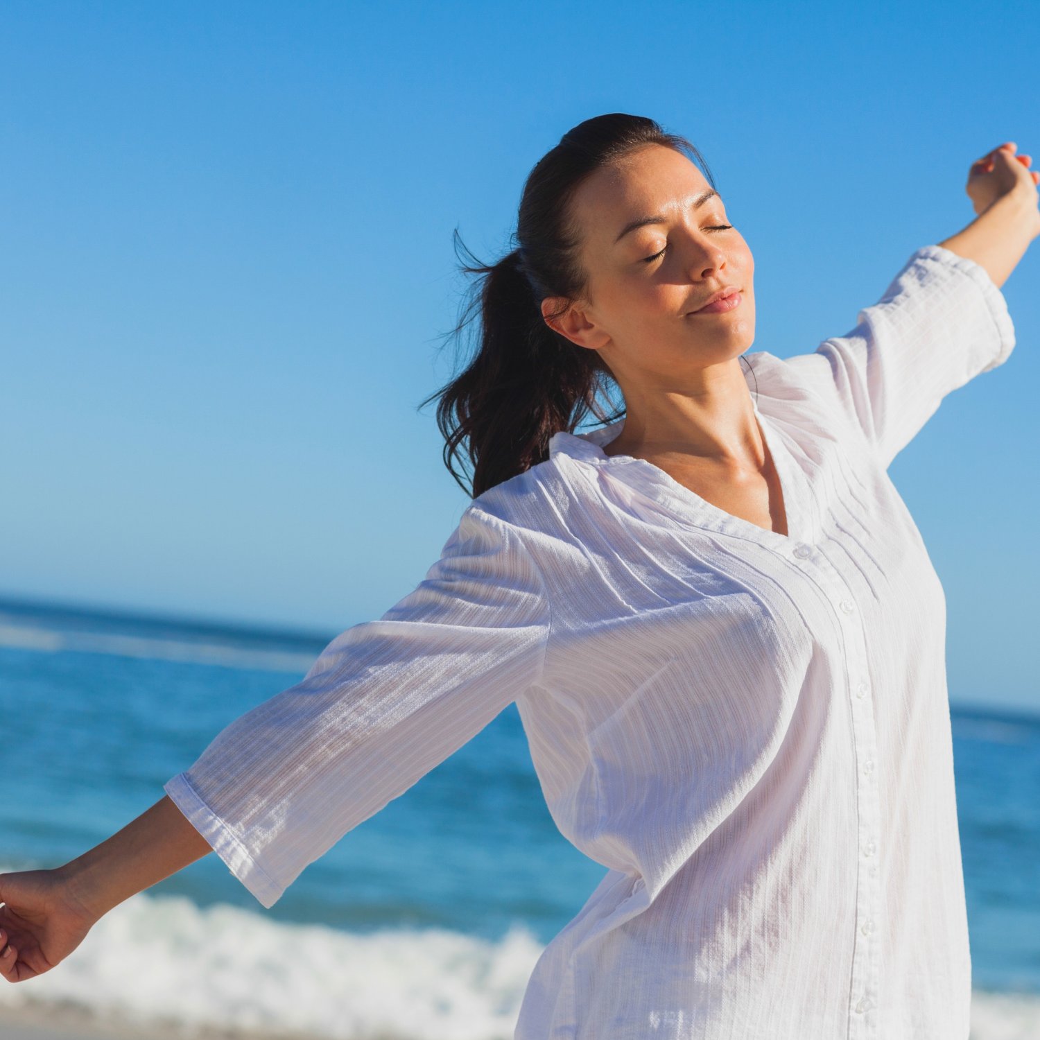 Woman standing by the ocean with arms outstretched and eyes closed, representing calmness, freedom, nervous system balance, healing, mindfulness, relaxation, and overall wellness.